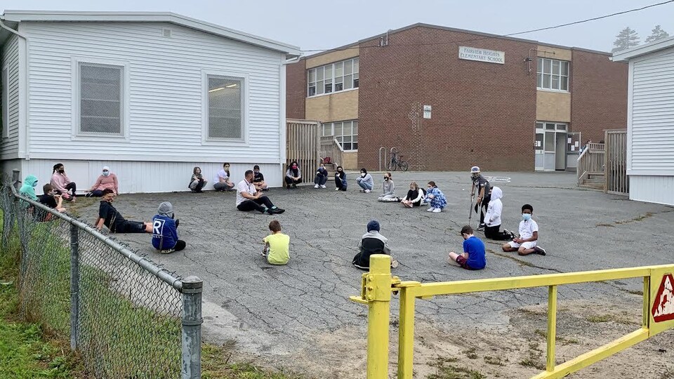 Des élèves et leur enseignants assis sur le sol derrière leur école.