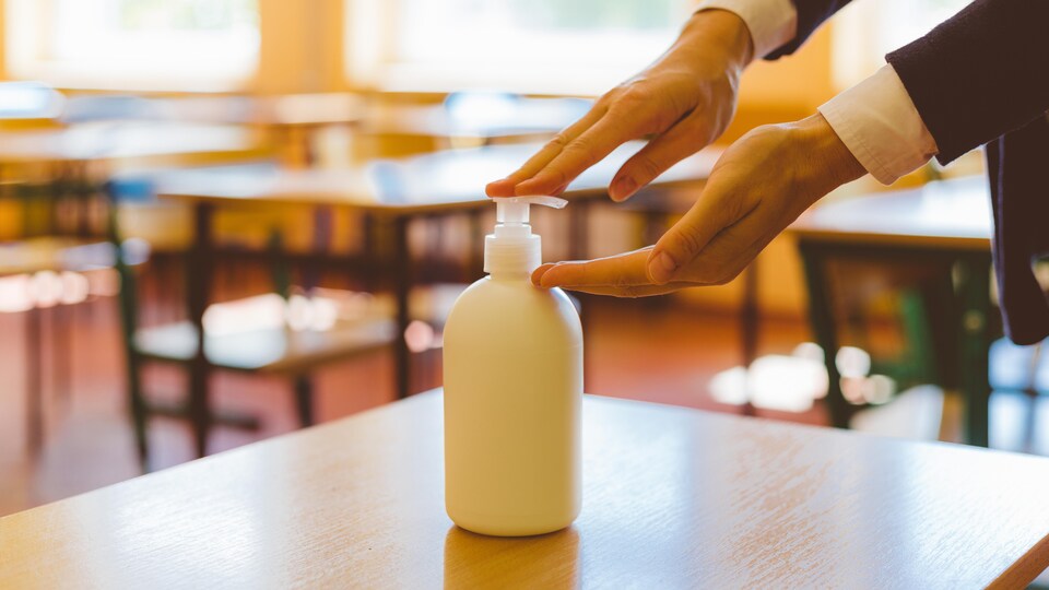 Les mains d'une enseignante qui appuie sur le bouchon d'une bouteille de désinfectant posé sur un pupitre dans une salle de classe.