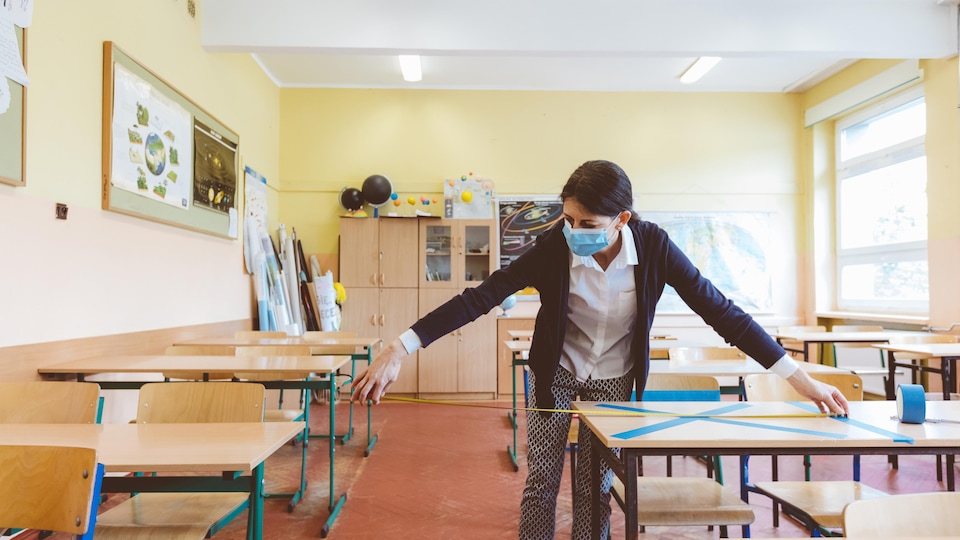 Une femme portant un masque bleu sur le visage mesure avec un mètre la distance entre deux pupitres dans une salle de classe.
