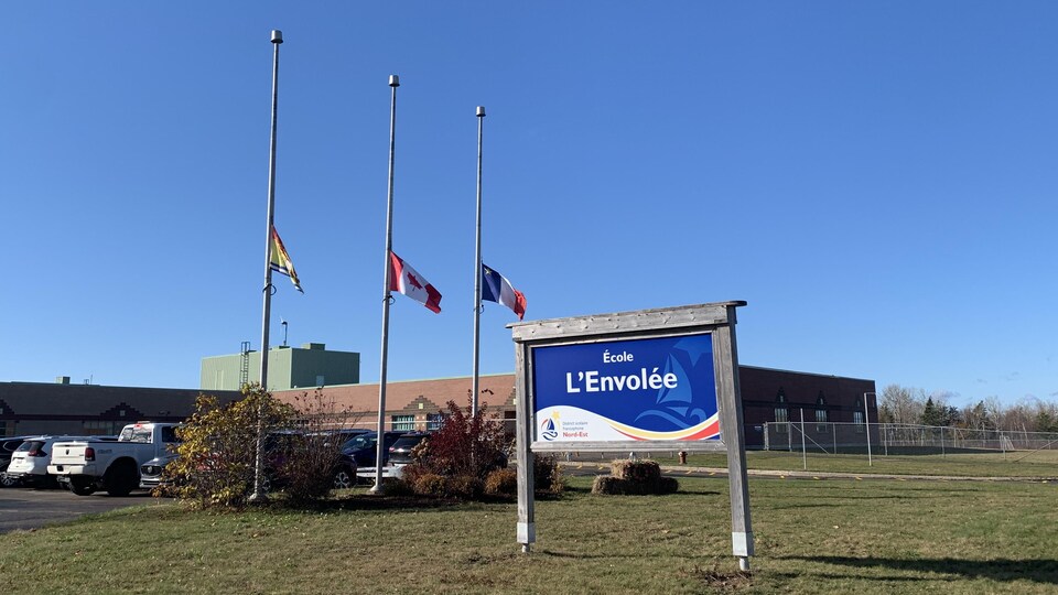 Un drapeau du Nouveau-Brunswick, un drapeau du Canada et un drapeau de l’Acadie sont en berne devant un établissement scolaire. Sur un panneau à l’entrée, il est écrit École L’Envolée.