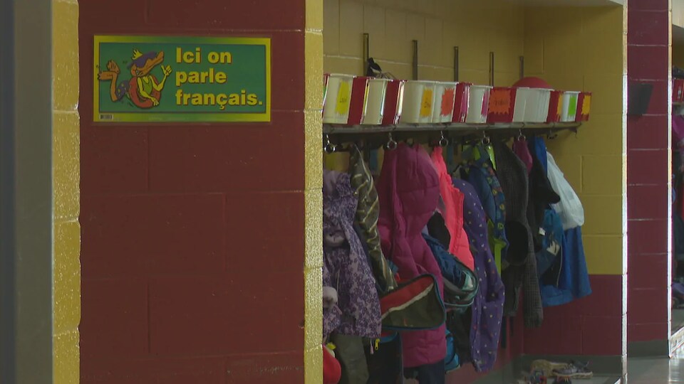 Des manteaux d'élèves dans un vestiaire d'une école. Une affiche indique « Ici, on parle français ».