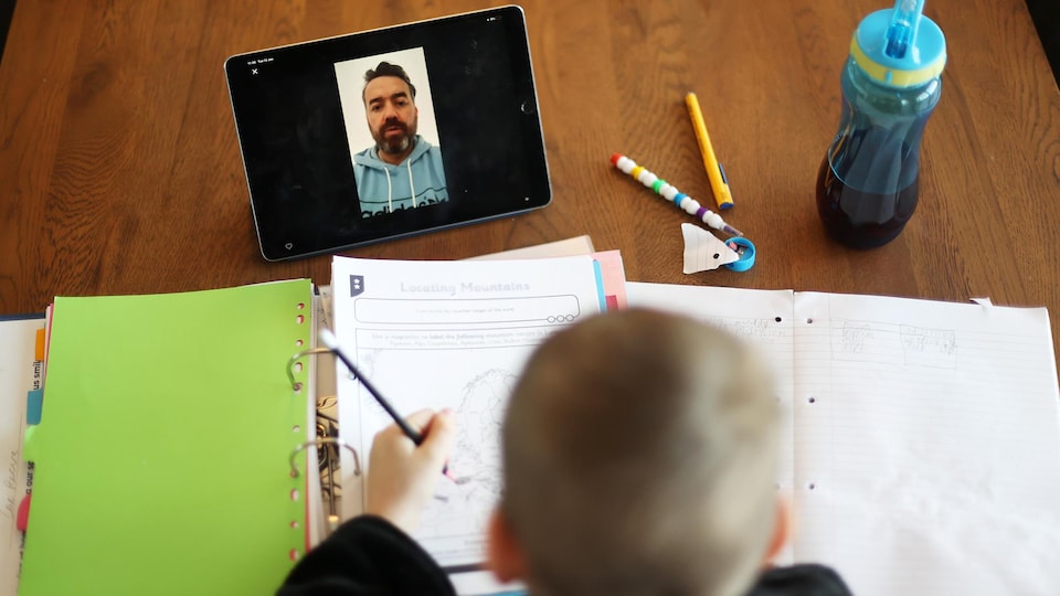 Un garçon photographié de haut devant ses cahiers d'école. Une tablette est posé devant lui et on y voit son professeur donnant le cours.