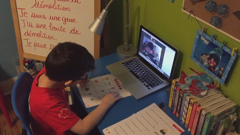Un enfant écrit sur une feuille placée devant un écran d'ordinateur sur lequel on voit un autre enfant faire la même chose.