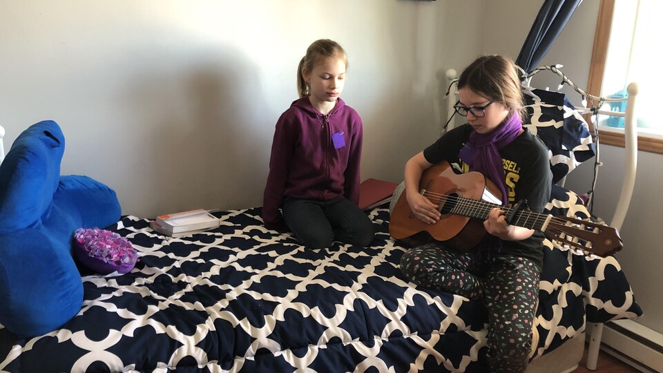Deux jeunes filles sont assises sur un lit alors que l'une joue de la guitare.