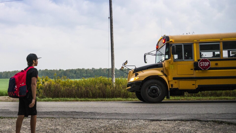 Un autobus scolaire s'immobilise sur une route rurale et un écolier marche sur l'accotement avec un sac à dos.