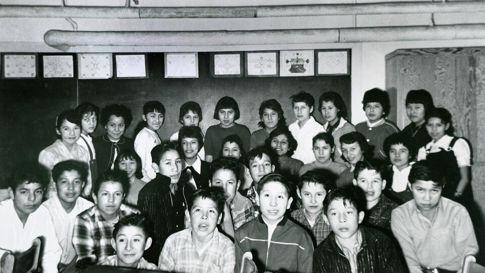 Des enfants posent pour la photo dans une classe. Ils sont debout, derrière une rangée de pupitres.