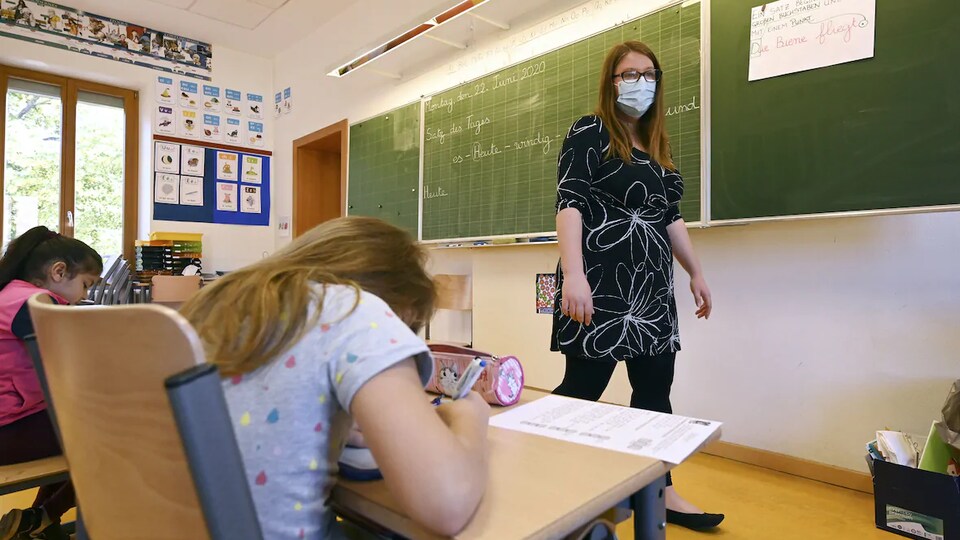 Une enseignante porte un masque. Elle se tient devant ses élèves, dans sa salle de classe..