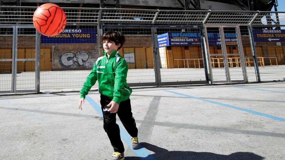 Un jeune garçon joue au ballon devant un stade protégé par un grillage.