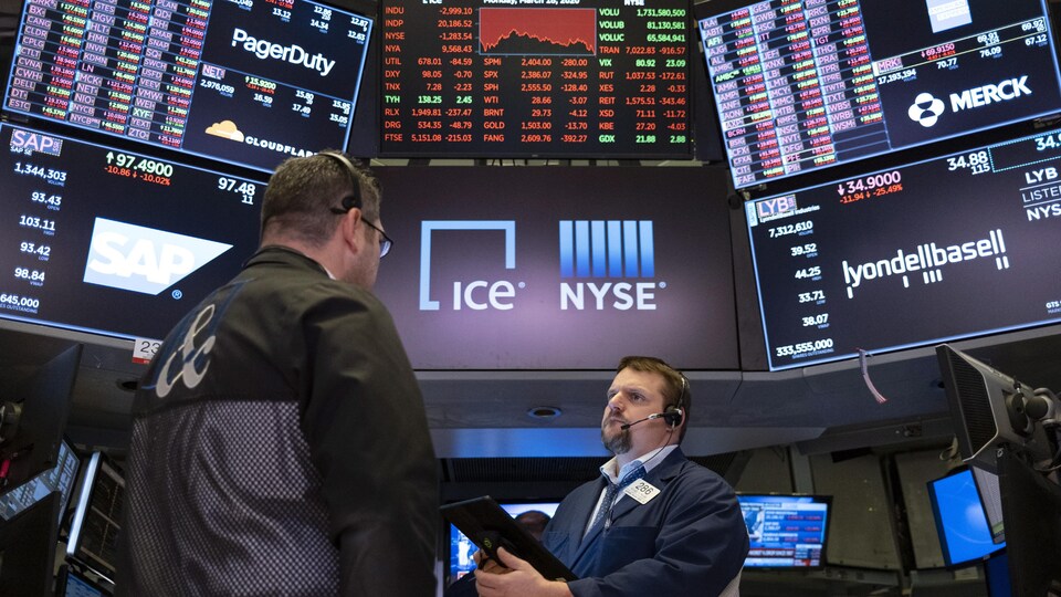 Deux hommes discutent devant de grands écrans sur lesquels s'affichent des données financières à la Bourse.