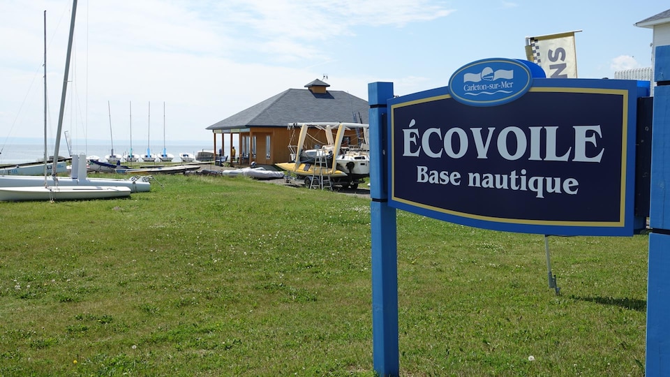 Une pancarte annonce la base nautique Écovoile. Derrière, on aperçoit un bâtiment de services ainsi que des bateaux.