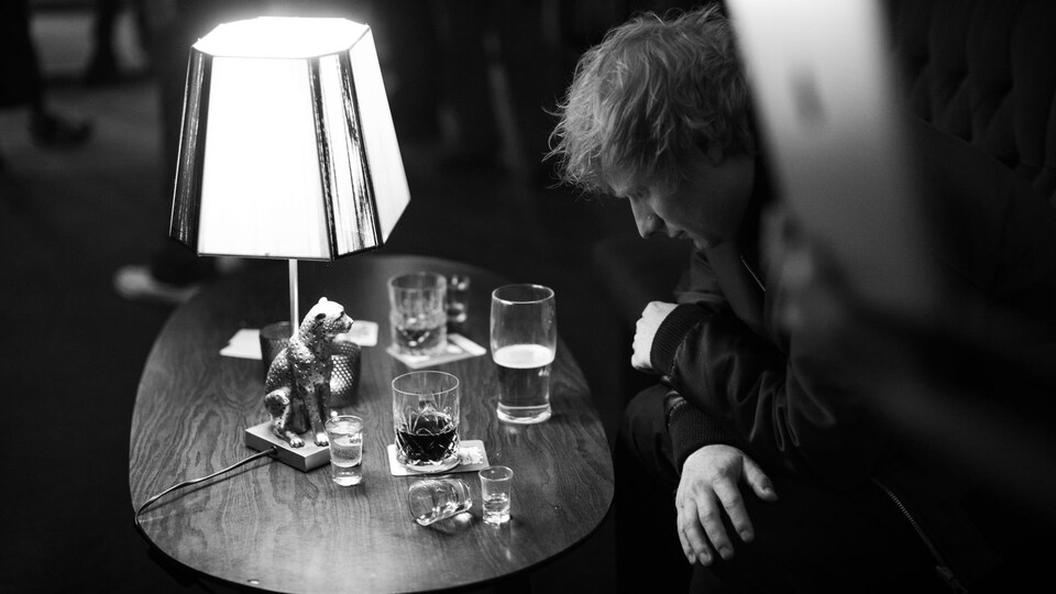 L'artiste est assis devant une table remplie de verres de bière et d'alcool; il regarde le sol, les bras croisés. 