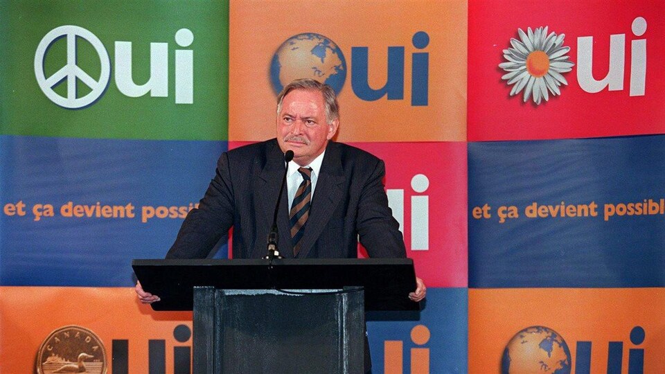 L'homme est debout sur une scène et parle dans un micro. Des affiches du oui du référendum sont en arrière de lui.