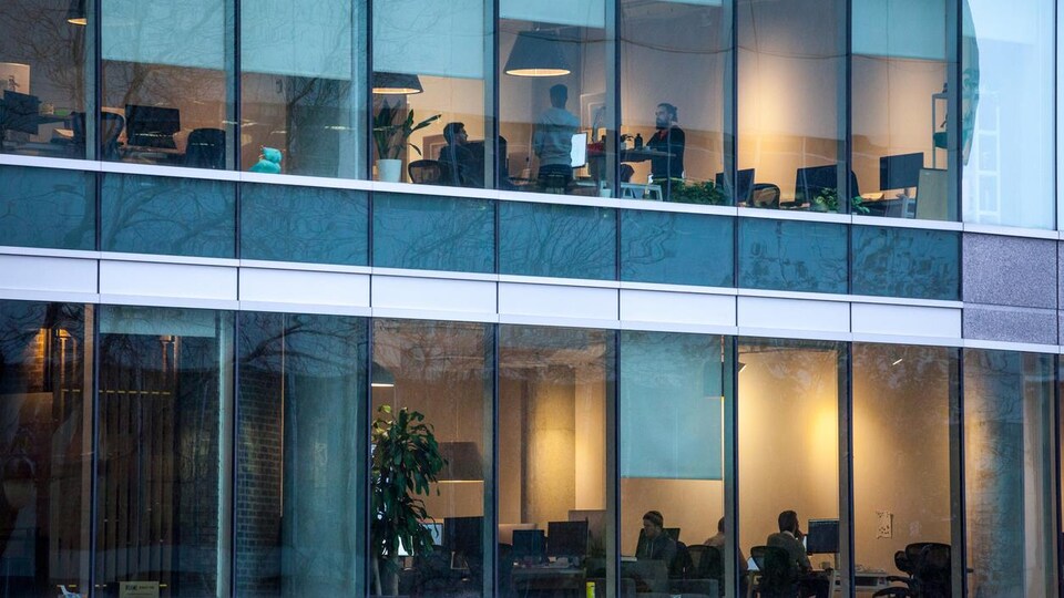 À travers la façade vitrée d'un bâtiment où se trouvent des bureaux, des gens travaillent.