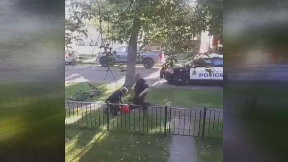 Un policier se dirige vers un homme maintenu au sol par un autre agent, dans une rue résidentielle.
