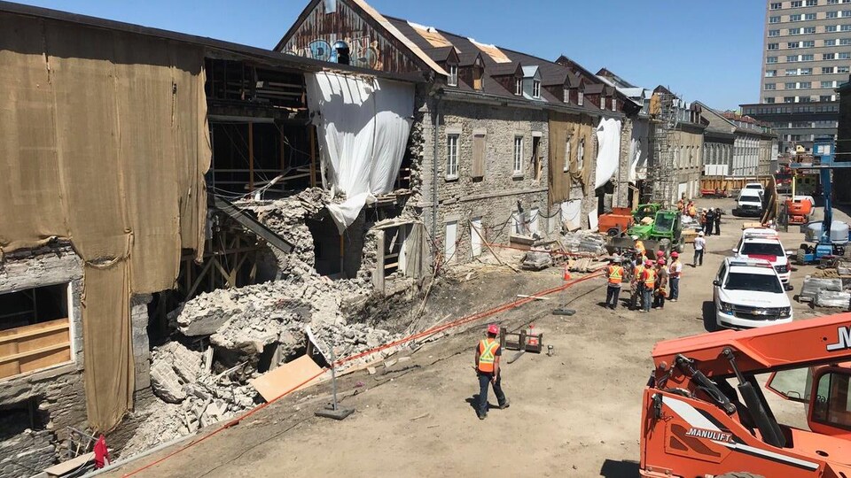 Vue sur le mur de pierre qui s’est effondré. On aperçoit des travailleurs et des véhicules d’urgence.