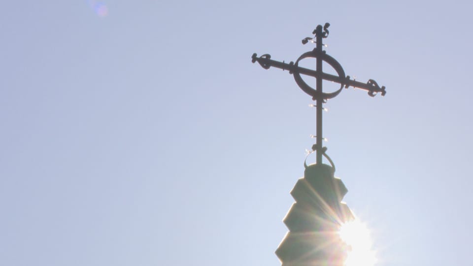 Une photo à contre-jour d'une croix au sommet d'une église. Le soleil brille derrière. Le ciel est bleu.