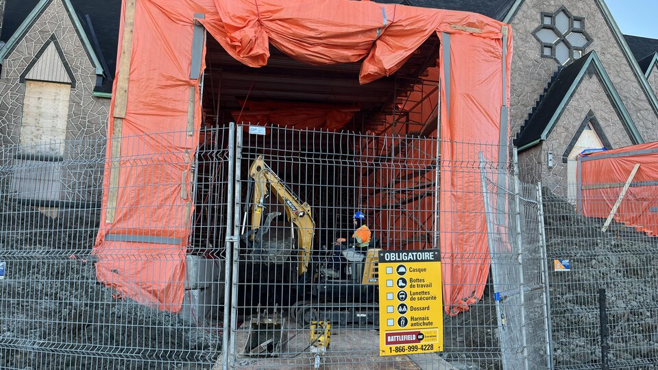 Un travailleur manipule une pelle mécanique sous une toile orange derrière une clôture de chantier.