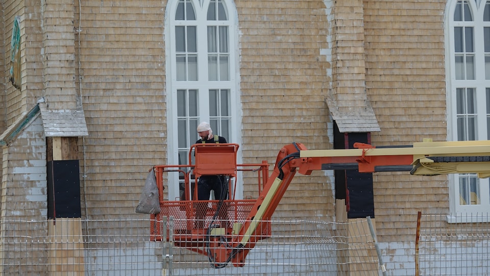 Un travailleur dans une nacelle devant un mur de bardeaux décapés.