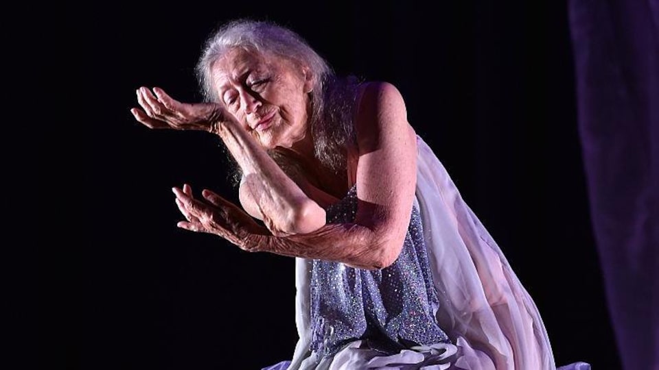 Une femme âgée, vêtue d'une robe mauve, danse sur scène.