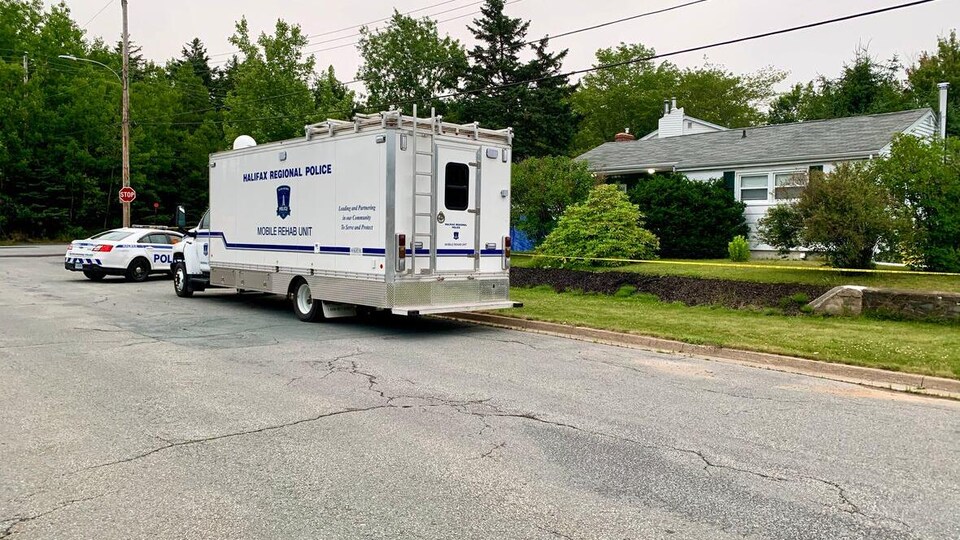Un camion et un véhicule de police sont stationnés devant une maison cachée derrière des buissons.