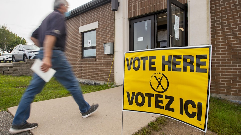 Un homme marche à grands pas vers la porte d'entrée d'un édifice à côté duquel est plantée une affiche jaune qui dit «Votez ici».