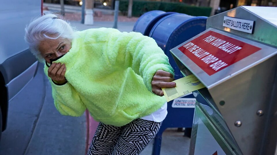 Une femme âgée se cache le nez et la bouche dans le col de sa polaire en déposant son bulletin de vote dans une boîte prévue à cet effet.