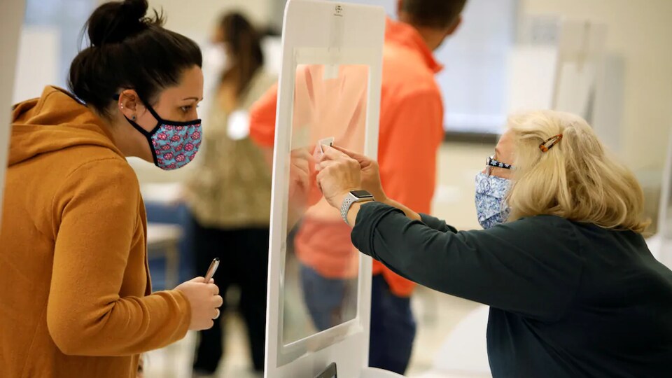 Une femme montre un bout de papier à une électrice à travers une barrière de protection translucide.
