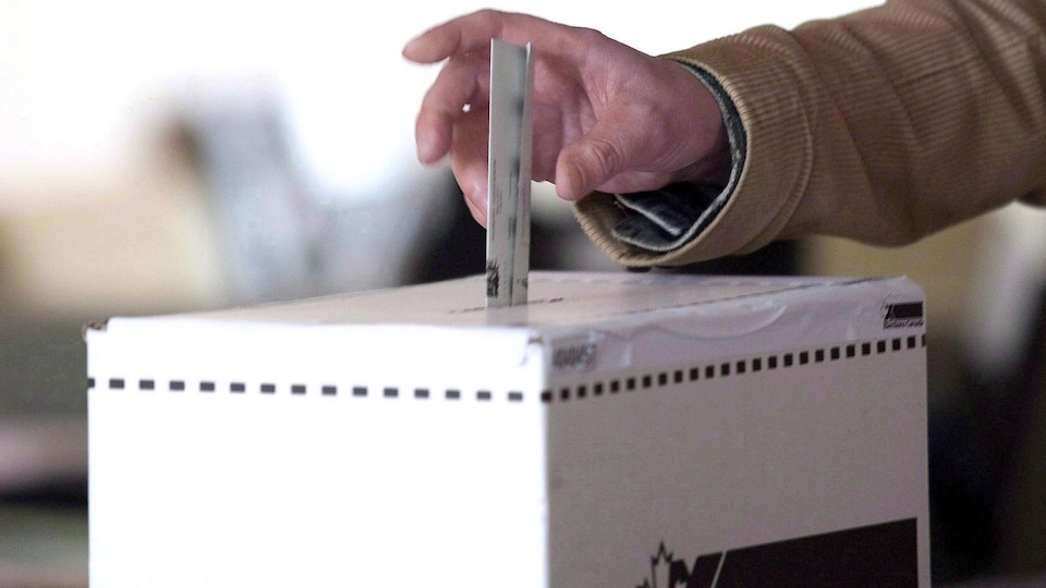 une main tient un bulletin de vote et la dépose dans une urne électorale. 