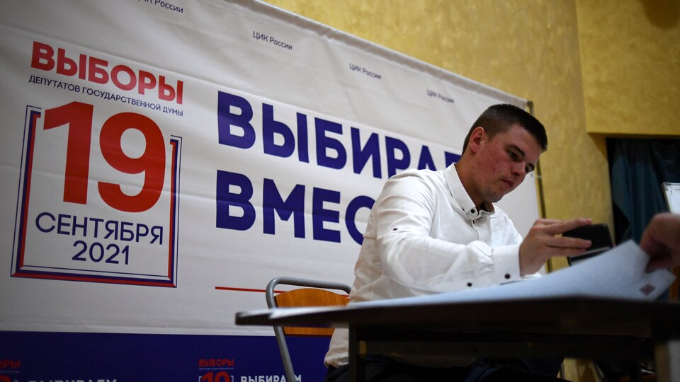 Un membre d'un bureau électoral russe regarde une série de documents sur un bureau, avec une affiche électorale derrière lui.