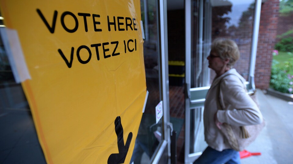 Un panneau est affiché sur la porte d'entrée du bureau de vote avec une inscription bilingue : Vote here, votez ici. Une femme entre par la porte.