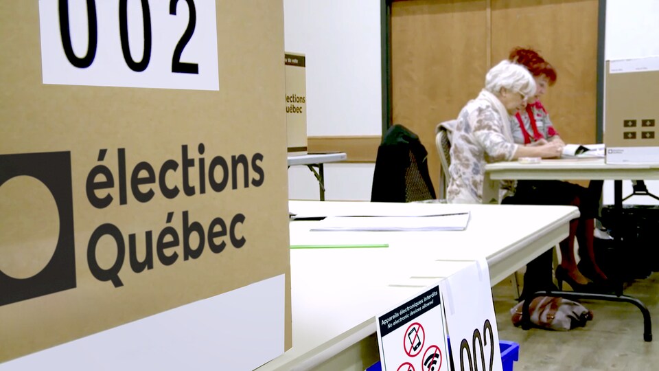 Deux femmes assises à un bureau sur lequel est déposée une urne électorale.