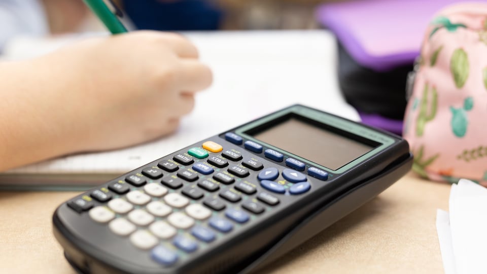 Un élève écrit sur une feuille à côté d'une calculatrice.