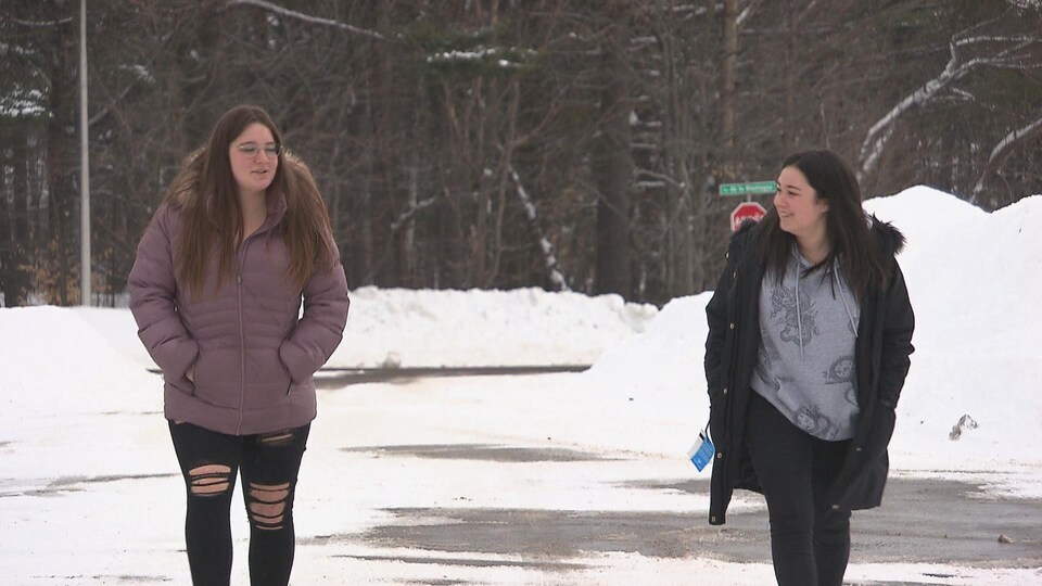 Deux jeunes femmes marchent à deux mètres de distance. Une porte un manteau mauve et l'autre a un manteau noir entrouvert.