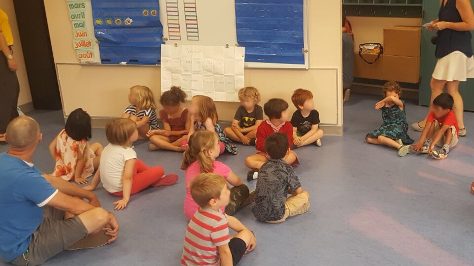 Des enfants assis par terre dans une classe de maternelle avec quelques parents en périphérie.