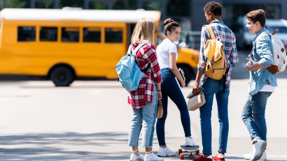 Des élèves du secondaire devant un autobus scolaire dans une cour d'école.