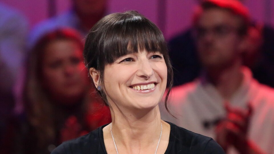 Elle sourit. Ses cheveux sont bruns. Elle porte un robe noire et un pendentif en argent.