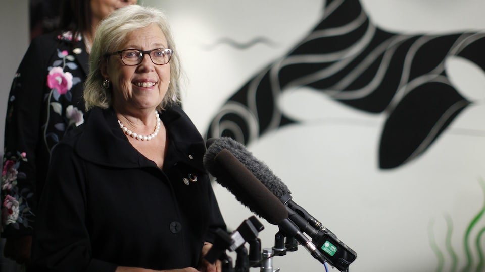 Elizabeth May sourit. Devant elle, plusieurs micros sont installés.