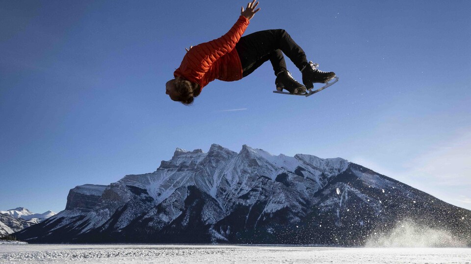 Elladj Baldé dans les airs, la tête en bas, en train de réaliser un saut périlleux arrière. Il est au-dessus d'un lac gelé.