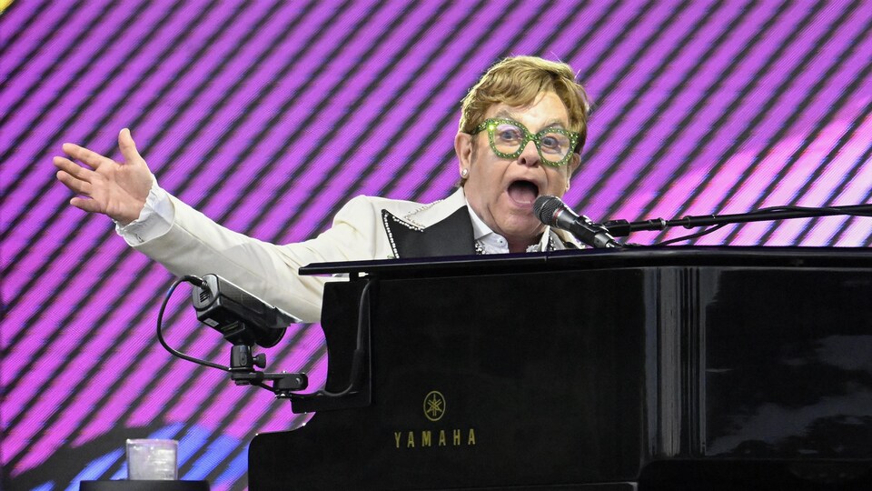 Elton John avec des lunettes vertes, un complet blanc, et un fond rose.