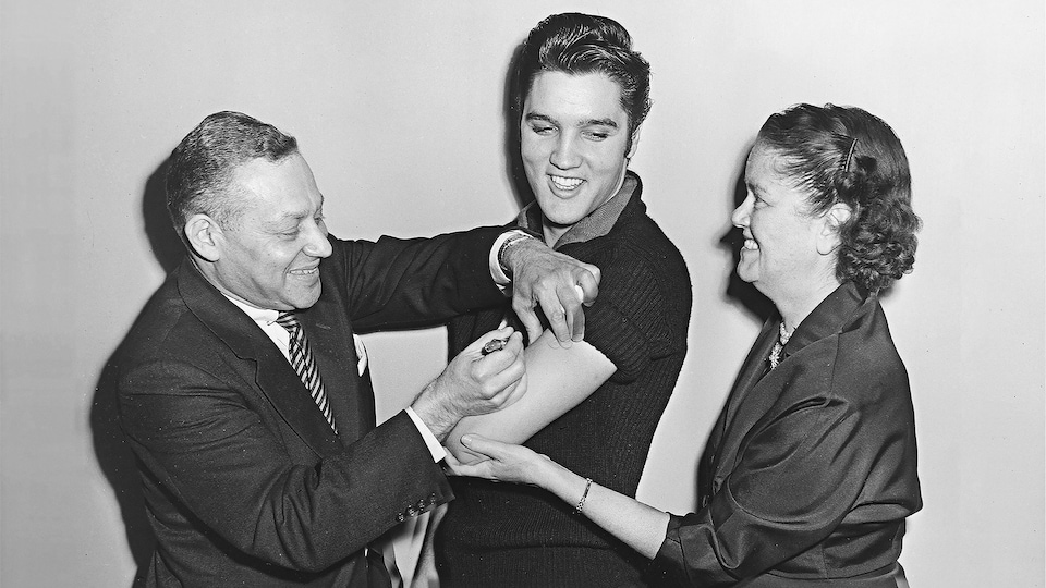 Elvis Presley, souriant, la manche retroussée, se fait injecter une dose de vaccin par un homme, à gauche, pendant qu'une femme, à droite, lui tient le bras.