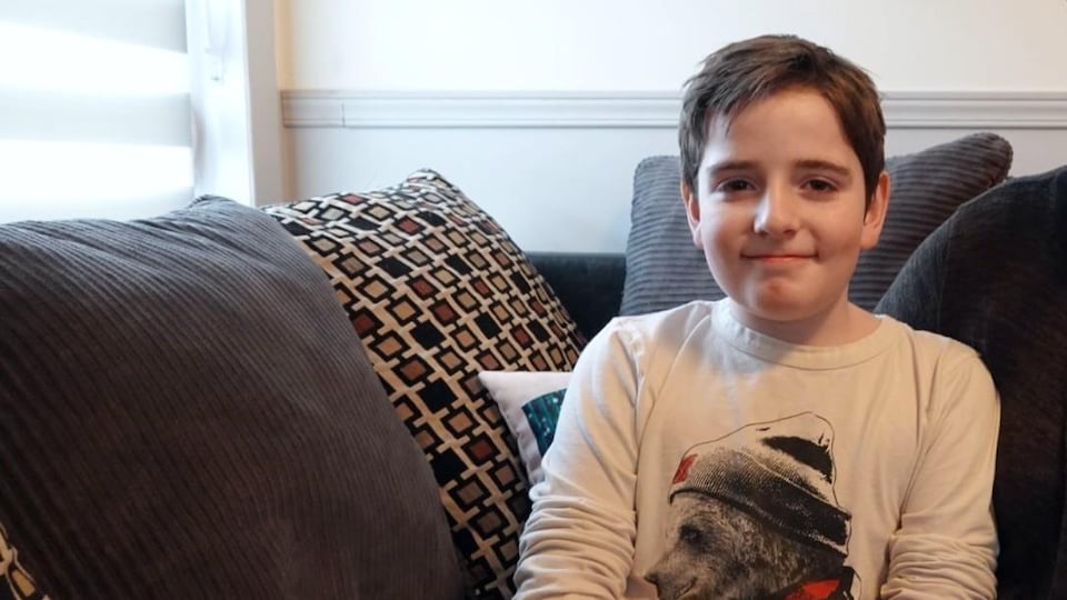 Elwan est assis sur un divan. Il porte un chandail avec un gros ours dessus. Il sourit.
