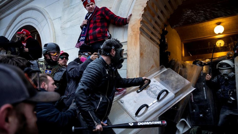 Des manifestants tentent de faire intrusion dans le Capitole, usant de violence pour pénétrer dans l'édifice, que tentent de défendre des policiers. À l'avant-plan, un émeutier portant un masque à gaz et armé d'un bâton de baseball et d'un bouclier.