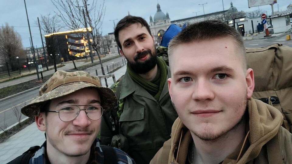 Trois hommes en treillis militaire prennent un égoportrait ensemble à l'extérieur.