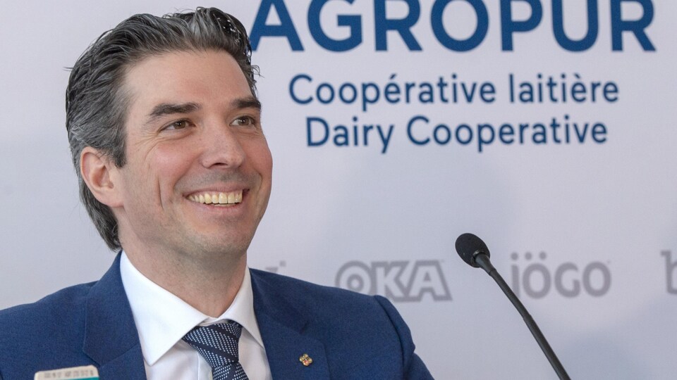 Émile Cordeau, souriant, en gros plan, devant une affiche sur laquelle il est écrit : Agropur, coopérative laitière.