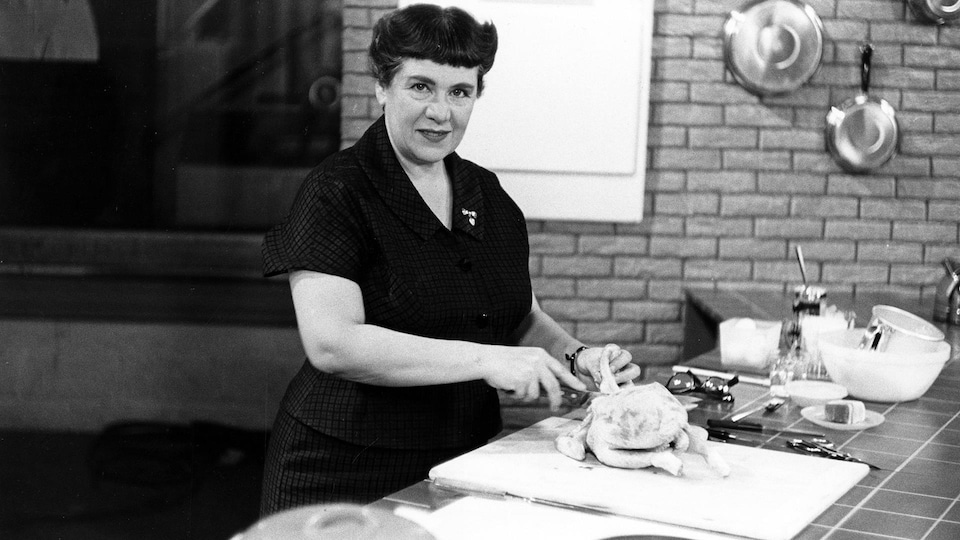 Dans une cuisine, la chroniqueuse Jehane Benoît découpe un poulet cru sur une planche.