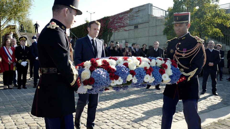 Emmanuel Macron est derrière une couronne de fleurs tenue par deux hommes en uniforme.