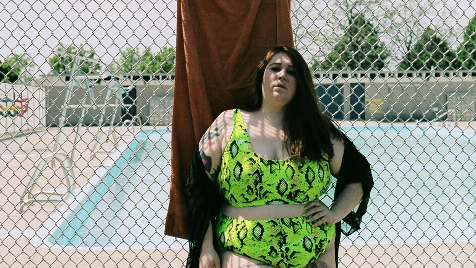 Une jeune femme pose en maillot deux pièces, vert fluo, devant la clôture métallique d'une piscine publique.