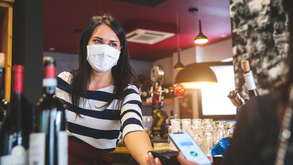 Une employée d'un restaurant, le visage couvert d'un masque, tend la machine interac à une cliente qui va payer avec son téléphone cellulaire.