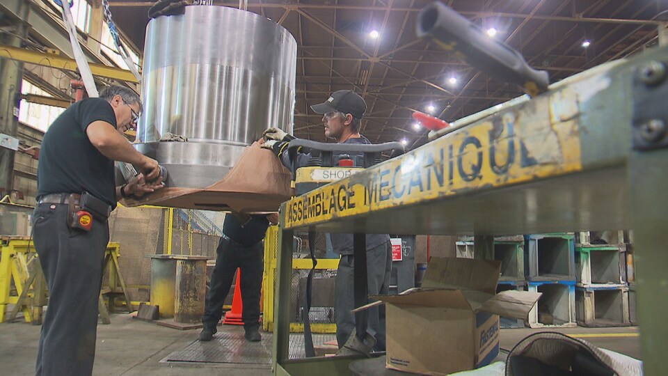 Des travailleurs manipulent une pièce mécanique suspendue dans une usine.