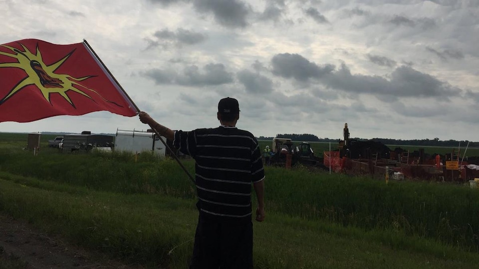 Un homme de dos brandit un drapeau face à des travailleurs affairés à la construction d'un pipeline.